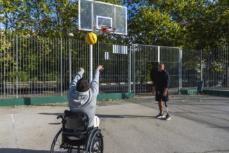 Two young men playing basketball outdoors, one in a wheelchair shooting the ball towards the hoop