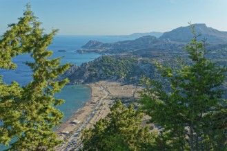 View of the Tsambika sandy beach, with mountains and blue sea, framed by green trees, Church of the