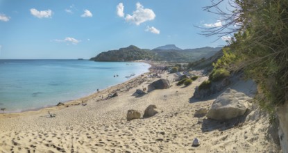 A sunny Tsambika sandy beach with clear blue water and few clouds in the sky, surrounded by rocks,