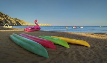 Colourful kayaks lying on Tsambika sandy beach, next to a pink flamingo pedal boat, boats floating