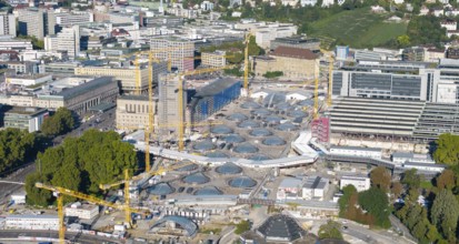 View of an urban construction site with cranes and modern glass structures, surrounded by green