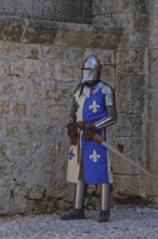 A knight in medieval armour stands with a sword in front of a stone wall, Medieval Town of Rhodes,