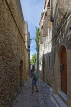 A woman with a rucksack walks through a narrow cobbled alley between historic stone houses,