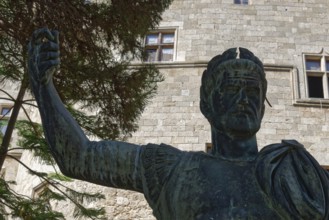 Statue of a man in ancient armour in front of a stone building with windows and trees, Grand