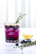 Refreshing blueberry and thyme drink served with lemon in a clear glass on a light surface