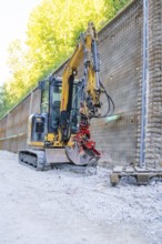 Mini excavator next to a metal mesh construction wall on a building site, Hermann Hessebahn