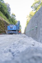 Blue excavator on a path next to a concrete wall with green pipes, Hermann Hessebahn construction