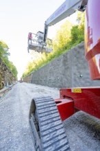 Red lifting platform on a gravel path in front of a concrete wall in the sunshine, Hermann