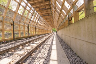 Light-flooded wooden tunnel with railway tracks and concrete path, surrounded by nature,