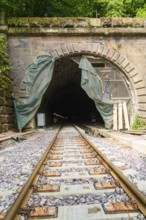 Railway tunnel with rails and stone wall, surrounded by green plants, renovation work in progress,