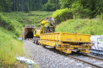 Yellow construction vehicle with equipment near a railway tunnel in green surroundings,