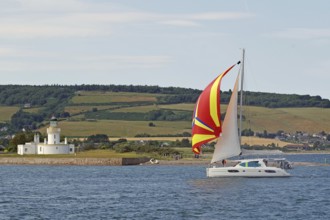 A sailing boat with a colourful sail sails near a lighthouse on the coast, Moray Firth, Inverness,