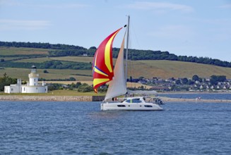 A sailing boat with a coloured sail sails near a lighthouse in front of a green landscape, Moray