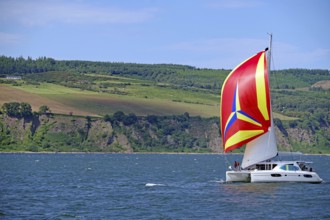 A sailing boat with a vividly coloured flag sails in front of a mountainous coastal landscape,