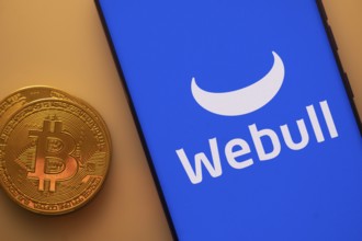 Bitcoin coin next to a smartphone showing Webull logo on a blue background