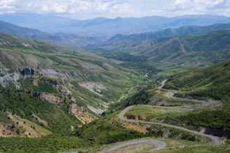 Winding roads lead down through a green valley into mountainous terrain under a clear sky, view