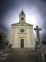 Yellow church with tower and cross in a cemetery under a cloudy sky, Kammersdorf Hollabrunn