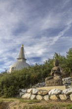 Buddha statue sitting on stones with a pagoda in the background and cloudy sky, Stupa am Wagram