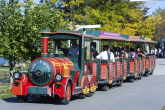 A colourful miniature train with cheerful tourists travels along a tree-lined road, Tulln