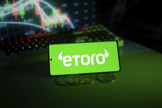 EToro logo on phone with coins and stock chart backdrop, evoking digital trading atmosphere