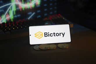Bictory logo on smartphone amidst coins and stock chart, conveying digital and financial themes