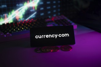 Currency.com logo on phone, with coins and stock chart suggesting financial trading platform