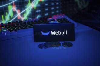 Webull depicted on a smartphone with coins and stock market indicators