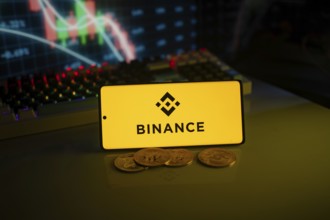 Yellow Binance logo with cryptocurrency coins on a desk, trading graphs in the backdrop