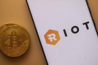 Bitcoin coin near phone displaying RIOT logo on a white background