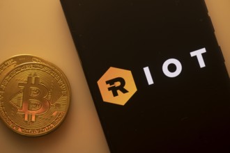 Bitcoin coin next to phone showing Riot Blockchain logo with black and hexagonal design