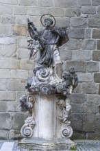 Stone statue of a saint on an ornate pedestal, baroque details in front of a stone wall, parish