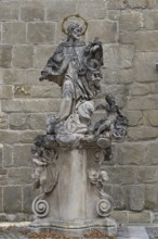 Baroque stone statue of a saint with angel on a decorated pedestal in front of a stone wall, parish