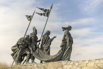 Sculpture group of historical figures with flags on a stone platform under a blue sky,