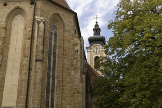 Gothic church with tower and clock next to green trees and bright sky, parish church St.Stephan