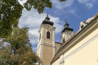 Baroque church towers with clocks in front of a blue sky, surrounded by autumnal trees, parish