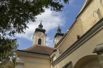 Two baroque church towers with clocks rise into a clear blue sky, surrounded by trees,