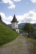 A clock tower at the end of a cobbled path, surrounded by green nature and mountains, Bruck an der