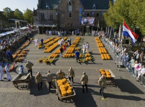 Participants on a market square with cheese wheels and flags, surrounded by people in a festive