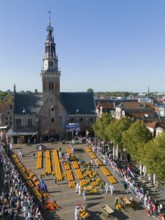 Market square with cheese wheels in front of a church and many visitors in sunny weather, aerial