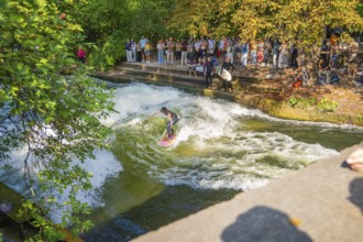 A surfer navigates through a river wave while spectators watch on the shore, Munich, Germany