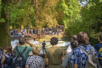 A group of people watching surfers riding in a river, surrounded by trees and summer nature,