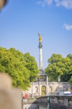 Column statue in golden spire, surrounded by green nature and bright blue sky, Munich, Germany