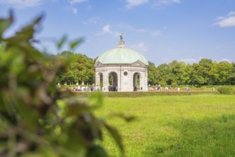 Meadow environment around grey pavilion with green dome, clothed by resting people and blue sky,