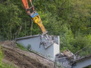 An excavator destroys a concrete wall on a slope, remains of a ladder visible, bridge demolition,