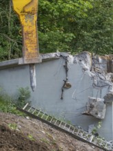 A demolition hammer destroys concrete, a ladder leans against the wall, surrounded by plants,