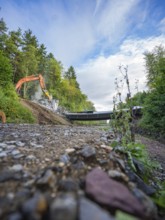 Wide angle shot of an excavator on a gravel road demolishing a bridge, surrounded by lush greenery