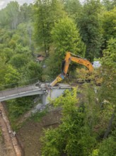 Excavator tears down part of a bridge in a green forest, an empty path runs underneath, bridge
