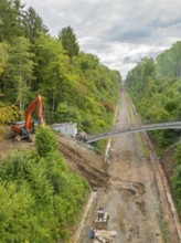 A single excavator during demolition work on a bridge section, next to a straight forest road,