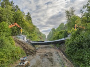 Excavators on both sides of a bridge during demolition work, surrounded by forest under a cloudy