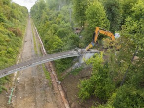 Bird's eye view of an excavator demolishing a bridge in the middle of the forest, with an empty